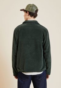 Veste en velours côtelé vert avec une surface texturée, manches longues et coupe décontractée, associée à un pantalon en denim foncé et une casquette bicolore.