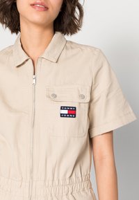 Béžový krátký overal z bavlněné směsi, vyznačující se předním zipem, elastickým pasem a nášivkou s logem na kapse na prsou.