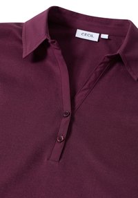 Burgunderrotes Poloshirt mit klassischem Kragen, zwei Knopfleiste und glatter Textur. Markenschild innen am Kragen sichtbar.