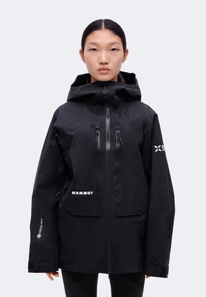 Une femme présente une veste Mammut noire à capuche, avec une fermeture éclair sur le devant, des poches poitrine et des logos sur les manches, sur un fond uni.