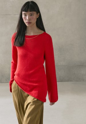 Giovane donna con capelli lunghi e scuri che indossa un maglione rosso asimmetrico e pantaloni marrone chiaro su sfondo grigio uniforme.