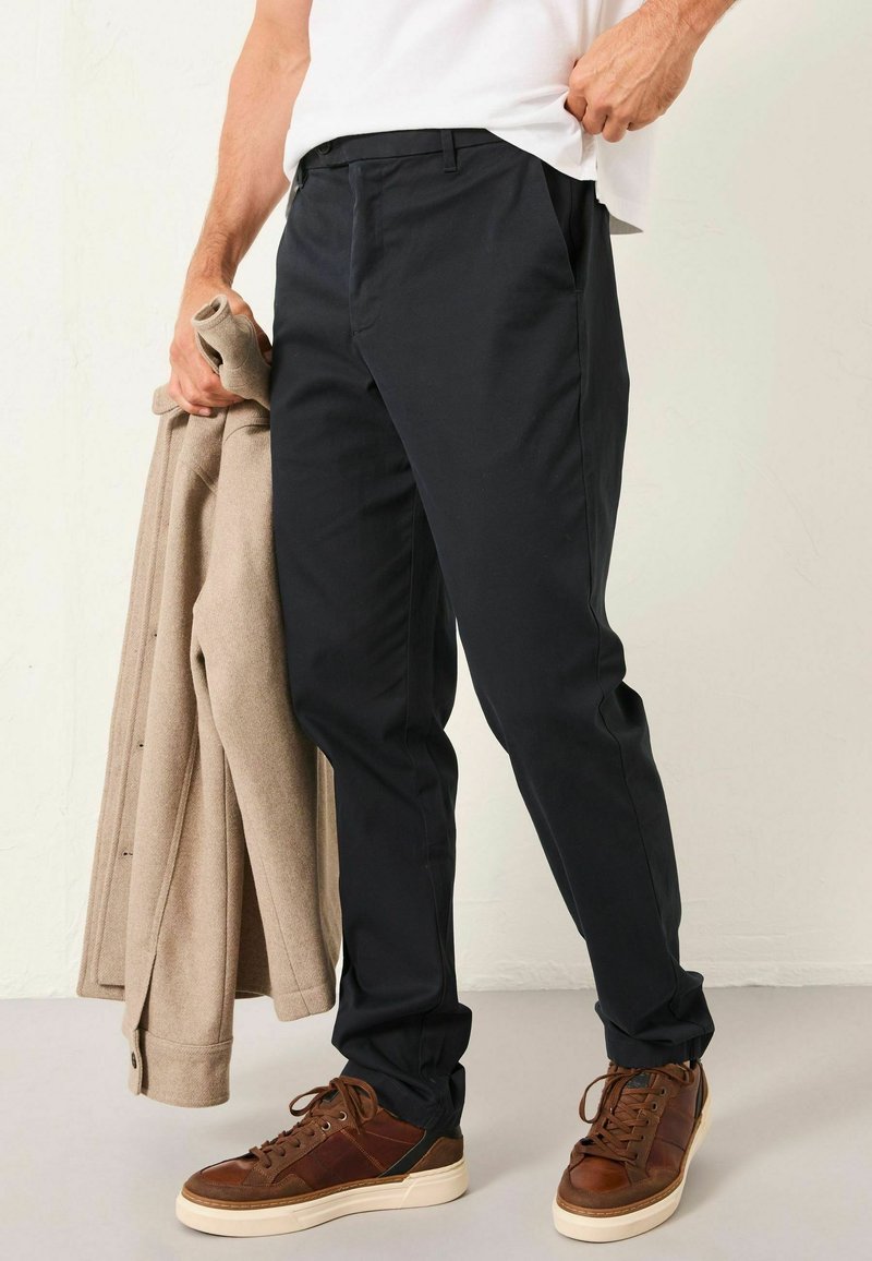 Marineblaue Hose mit tailliertem Schnitt, aus glattem Stoff. Kombiniert mit einem beigen Mantel und braunen Sneakers. Einfaches Design ohne sichtbare Muster.