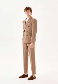 Costum cu două rânduri de butoni dintr-un material bej deschis, cu butoni închisi la culoare, pantaloni slim-fit și mocasini din suede maro, pe un fundal neutru.