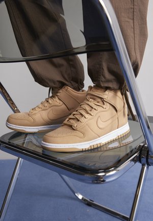 Baskets montantes beige avec semelles blanches portées par une personne debout sur une chaise transparente aux pieds chromés sur un sol bleu.