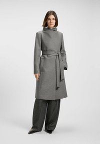Cappotto grigio in lana con colletto alto, vita regolabile con cintura e due tasche frontali. Indossato sopra pantaloni grigi a gamba larga e scarpe nere con punta.