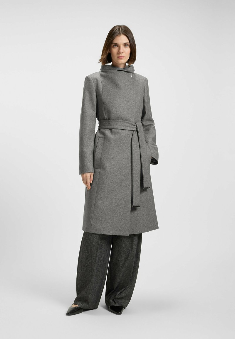 Cappotto grigio in lana con colletto alto, vita regolabile con cintura e due tasche frontali. Indossato sopra pantaloni grigi a gamba larga e scarpe nere con punta.