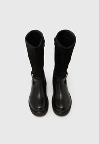 Črni škornji do sredine meč z usnjenim podplatom in semišasto gazo, opremljeni s stranskimi zadrgami in okrasnim trakom z zlatim sponkom na belem ozadju.