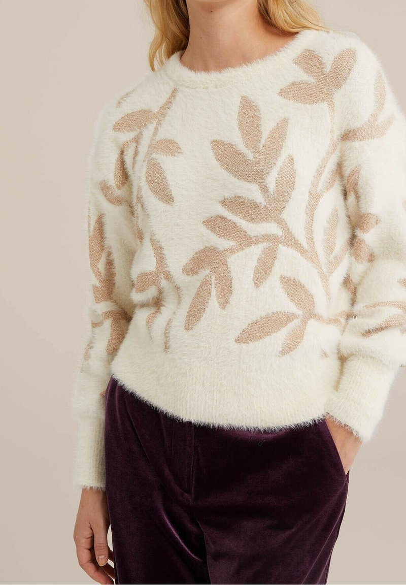 Femme portant un pull doux crème orné de motifs de feuilles beiges et un pantalon en velours violet foncé, posant avec une main dans la poche.