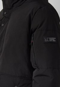 Schwarze Daunenjacke mit hohem Kragen, Druckknöpfen und einer Reißverschlusstasche am linken Ärmel. Enthält ein Markenpatch am Arm.
