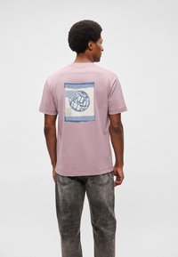 Απαλή ροζ βαμβακερή μπλούζα με κοντά μανίκια, που διαθέτει μπλε γραφικό σφαίρας και σχέδιο κειμένου στην πλάτη. Άνετη εφαρμογή και λαιμόκοψη τύπου crew.