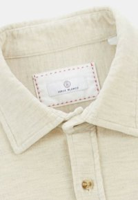 Chemise beige à boutons en tissu texturé, avec un col, une étiquette ronde portant "Serge Blanco" et des coutures rouges sur le bord de l'étiquette.