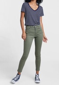 Jean skinny vert à taille haute, associé à un t-shirt à col en V rayé bleu marine et blanc. Porté avec des baskets montantes bleu foncé.