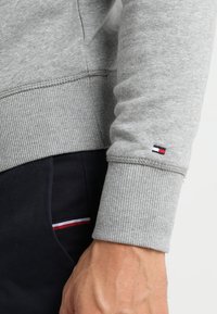 Hellgrauer, gerippter Sweatshirtbund mit kleinem rot-weiß-blauen Logo über dem Handgelenk, kombiniert mit schwarzen Hosen, die einen kleinen rot-weiß-blauen Streifen haben.