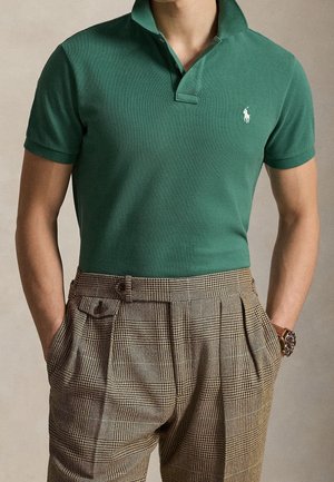 Homme portant un polo vert à manches courtes rentré dans un pantalon marron à carreaux taille haute, mains dans les poches et une montre-bracelet visible.