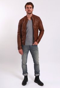 Braune Lederjacke mit gerippten Schultern, graues V-Ausschnitt-Shirt, hellblaue Jeans und schwarze Knöchelstiefel. Einfaches Design, taillierte Form.