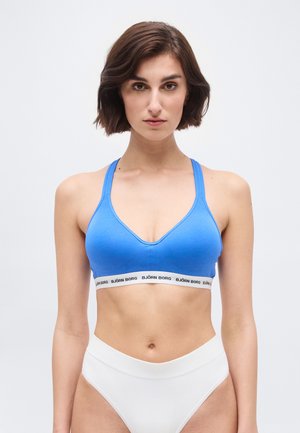 Frau mit kurzen braunen Haaren, die einen blauen Sport-BH und hoch taillierte weiße Unterwäsche vor einem schlichten weißen Hintergrund trägt.