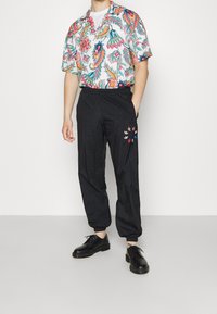 Lyhythihainen kukkakuvioinen paita, jossa on monivärisiä kuvioita, yhdistetty mustiin elastisiin housuihin, joissa on värikkäitä kukka-embroidauksia. Mustat kengät.