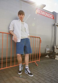 Lätt, randig skjorta med knappar över en krämfärgad t-shirt; denimshorts; svarta och blå höga sneakers; orange barriär i bakgrunden.