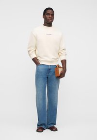 Άνδρας όρθιος, φορώντας κρεμ φούτερ και μπλε τζιν παντελόνι, κρατώντας μικρό καφέ ραδιόφωνο vintage, σε απλό φωτεινό φόντο.