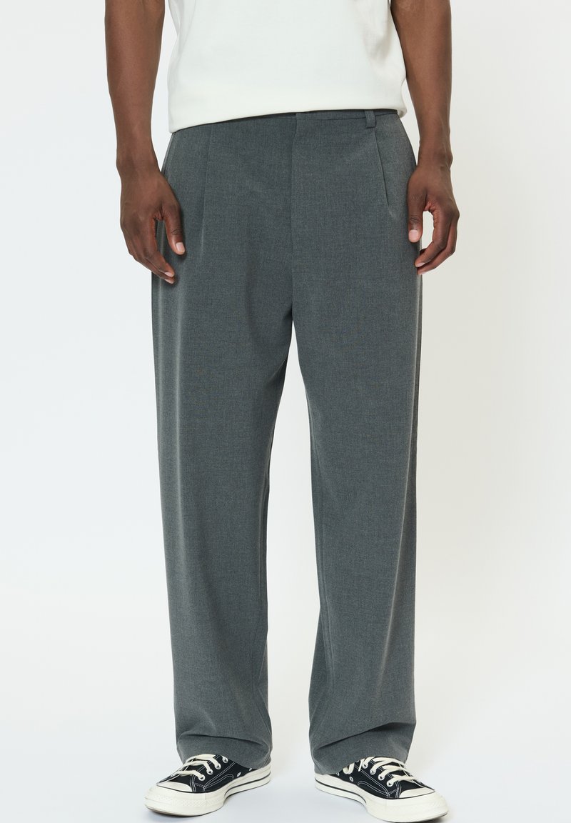 Sivé na mieru šité nohavice v úzkom strihu s záhybmi a hladkou textúrou, kombinované s čiernymi plátenými teniskami s bielymi šnúrkami.