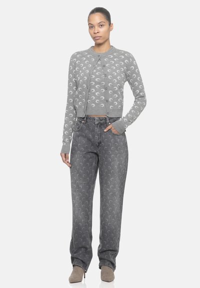 Donna in piedi con indosso un cardigan grigio abbinato a un top con motivi a mezzaluna, jeans grigi larghi con lo stesso motivo e stivaletti beige con tacco.