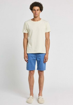 Jeune homme debout, portant un t-shirt beige uni, un short bleu et des chaussures beiges sans lacets sur un fond gris clair.