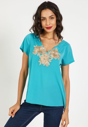 Mujer con cabello oscuro ondulado que lleva una blusa turquesa de manga corta con bordado floral beige y vaqueros azul oscuro, de pie frente a un fondo liso.