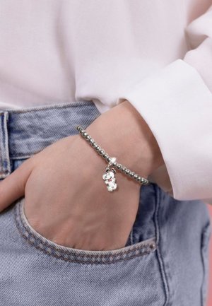 Polso con braccialetto in argento con perline che presenta un piccolo ciondolo Snoopy, mano infilata nella tasca di jeans blu, indossa una camicia bianca a maniche lunghe.