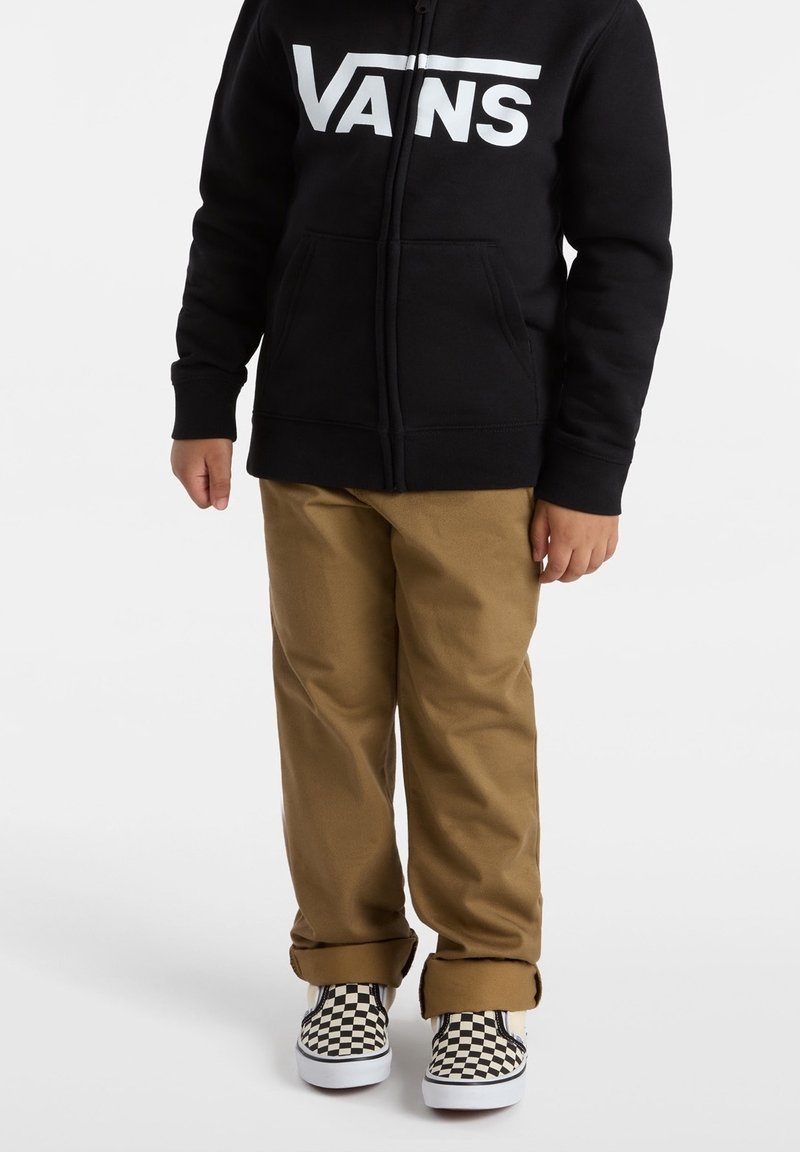 Enfant portant un sweat à capuche Vans noir, un pantalon beige avec les revers roulés, et des baskets slip-on à damier noir et blanc, debout sur un fond blanc.