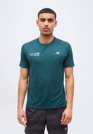 Νεαρός άνδρας φοράει πετρόλ κοντομάνικη μπλούζα με τα λογότυπα "tcs 20 26" και New Balance, συνδυασμένη με μαύρο σορτς με σχέδια, στέκεται μπροστά σε απλό φόντο.