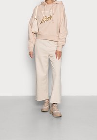 Felpa cropped rosa chiaro con lettering dorato, abbinata a pantaloni larghi color crema e sneaker beige. La texture è morbida e casual.