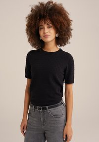 Melna īsām piedurknēm adīta jaka ar rakstu, apaļu kakla izgriezumu un ribotām piedurknēm. Apvienota ar augstā vidukļa pelēkiem džinsiem un melnu jostu.