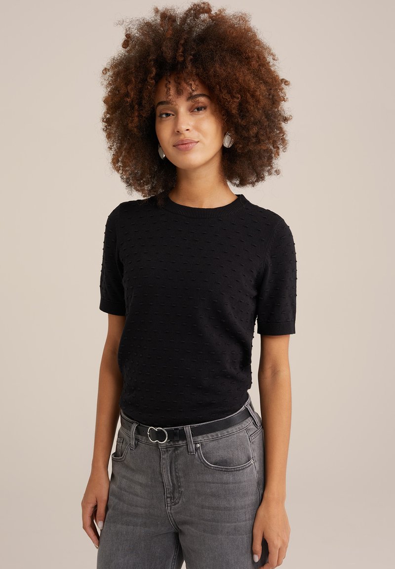 Melna īsām piedurknēm adīta jaka ar rakstu, apaļu kakla izgriezumu un ribotām piedurknēm. Apvienota ar augstā vidukļa pelēkiem džinsiem un melnu jostu.