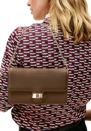 Brązowa skórzana torebka z złotym zapięciem, łańcuszkowym paskiem i prostokątnym kształtem, leżąca na wzorzystej czerwono-białej koszuli.