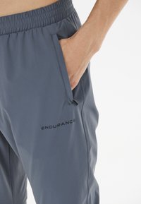 Graue Sporthose aus leichtem Material mit elastischem Bund und Seitentasche, mit schwarzem "ENDURANCE"-Logo.