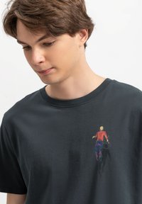 Zwarte T-shirt van katoen, met een kleurrafiek van een persoon in een rood en blauwe outfit, gepositioneerd aan de linkerkant.