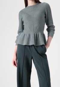 Femme portant un pull gris côtelé avec un ourlet à basque et un pantalon taille haute large gris foncé, debout avec une main dans la poche.