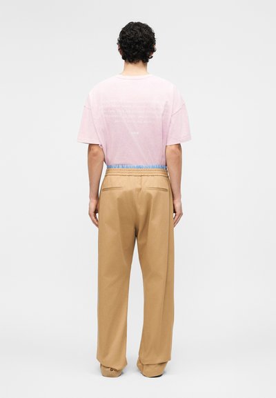 Άνδρας με σγουρά μαλλιά, στραμμένος προς τα πίσω, φοράει ροζ ανοιχτό κοντομάνικο πουκάμισο και μπεζ παντελόνι με φαρδιά μπατζάκια, ελαστική μέση και πίσω τσέπες.
