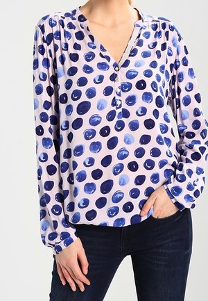 Hellviolette Bluse mit unterschiedlichen blauen Polka-Punkten, langen Ärmeln und Knöpfen am V-Ausschnitt, getragen zu dunklen Jeans.