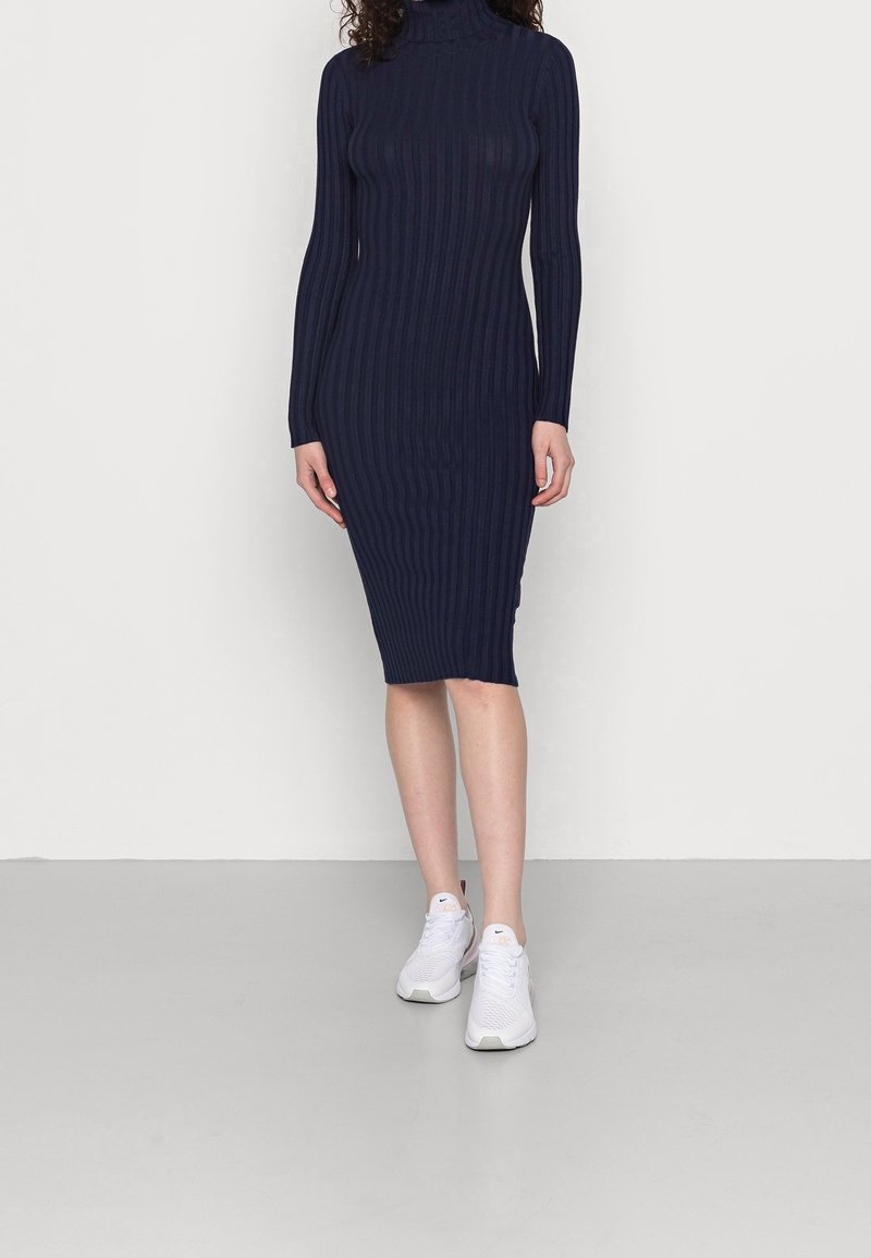 Femme portant une robe midi côtelée à col roulé bleu marine associée à des baskets blanches de sport, debout sur un sol et un mur gris clair unis.