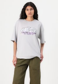 T-shirt gris oversized à manches courtes, avec un texte graphique coloré "ALPHA STUDIO EDITION" dans un design réfléchissant.