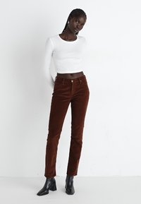 Bruine corduroy broek met een slim fit, voorzien van een sluiting met een knoop aan de voorkant en riemlussen. Gecombineerd met een witte shirt met lange mouwen en zwarte enkelboots.