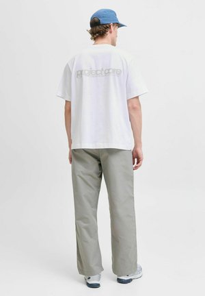 Weißes kurzärmliges T-Shirt mit einem grauen Logo auf dem Rücken, kombiniert mit lockeren grauen Hosen und einer hellblauen Cap.