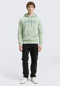 Felpa verde chiaro con tasca frontale e stampa del logo, abbinata a jeans neri e sneakers bianche. Texture morbida e vestibilità casual.