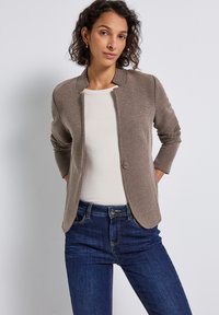 Blazer texturé marron clair avec un col rond, fermeture à un bouton et manches trois-quarts, associé à un haut beige et un jean foncé.