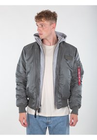 Giacca bomber grigia con design trapuntato, dotata di cappuccio grigio, polsini a coste e un patch con logo sul petto. Chiusura frontale con zip.