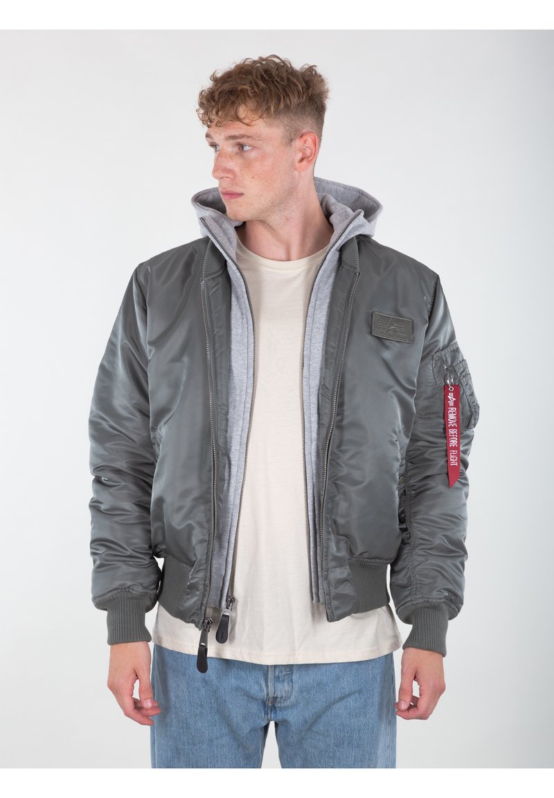 Giacca bomber grigia con design trapuntato, dotata di cappuccio grigio, polsini a coste e un patch con logo sul petto. Chiusura frontale con zip.