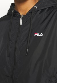 Svart huva jacka med frontdragkedja, med en liten vit och röd FILA-logotyp på vänster bröst och snoddar för huven.