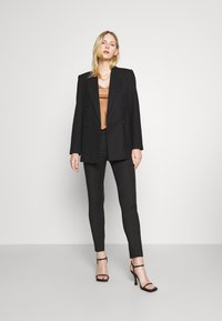 Blazer doppiopetto nero sopra un top bronzo, abbinato a pantaloni attillati neri e tacchi a strappo. Tessuto liscio e vestibilità sartoriale.