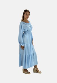 Lichtblauwe jurk met lange mouwen, een smock taille en een gelaagde rok, met een ronde halslijn en knoopdetails. Gedragen met sleehak sandalen.